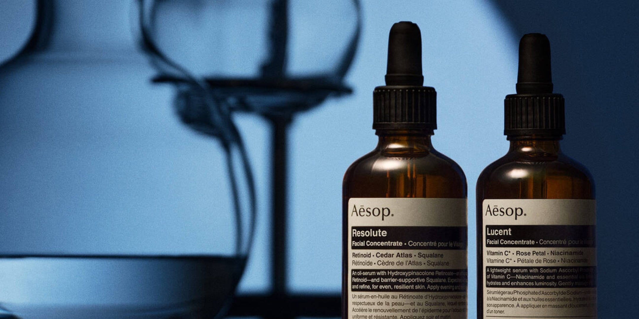 Two Aesop skincare dropper bottles, Resolute and Lucent, standing upright against a dark blue background with a blurred glass vessel.